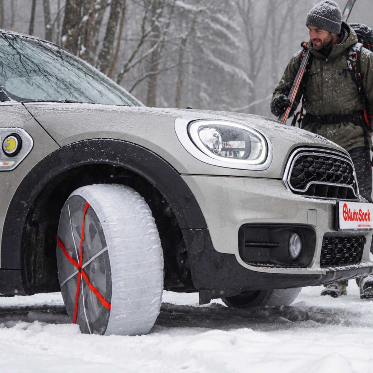 Mini Cooper argentata in un ambiente innevato, con una catena da neve montata sulla ruota anteriore. Sullo sfondo, un uomo con abbigliamento invernale e scarponi da sci si trova vicino all’auto.